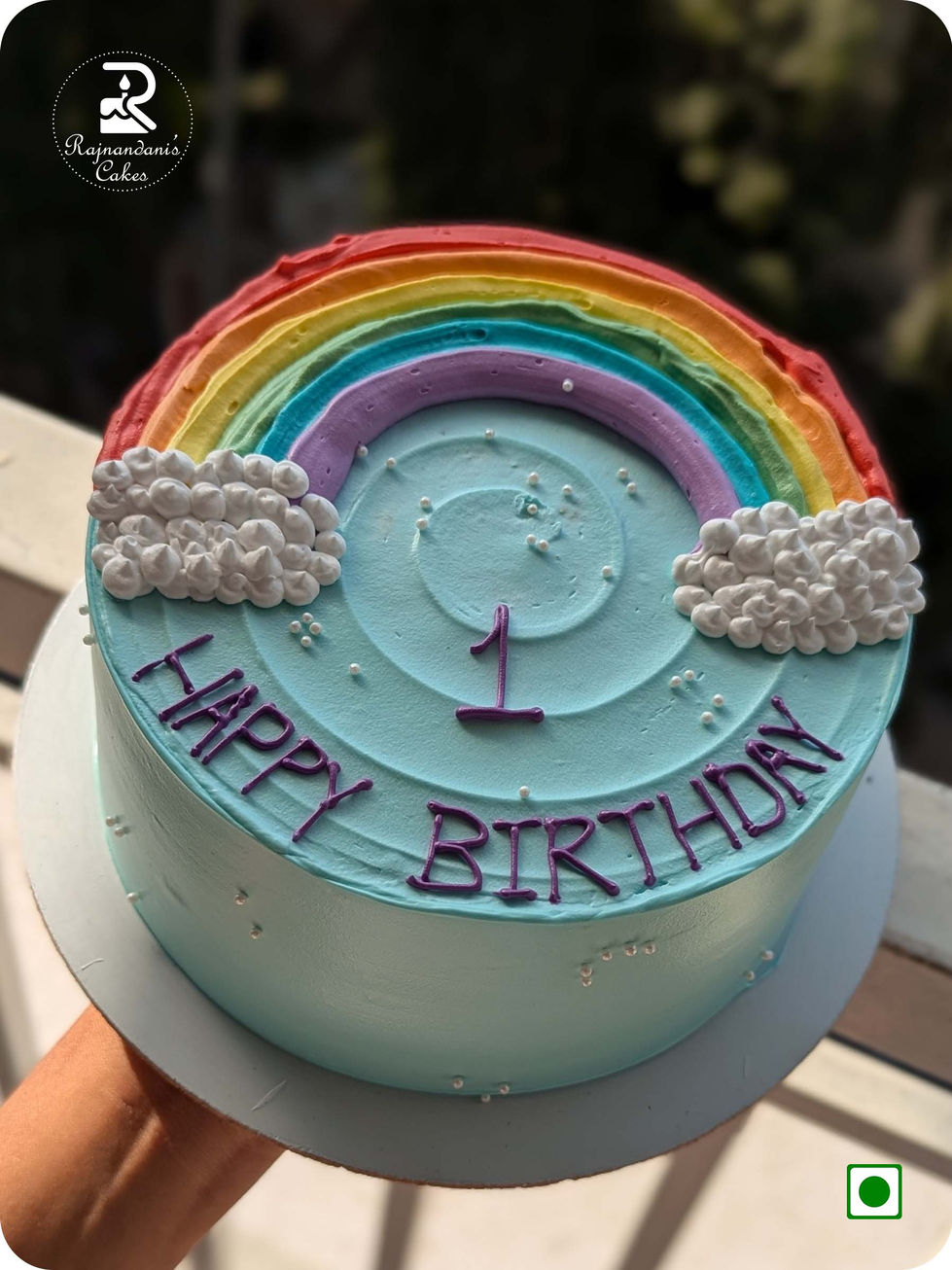 Rainbow first birthday cake featuring piped clouds, sugar pearls, and Happy Birthday lettering on sky blue frosting.