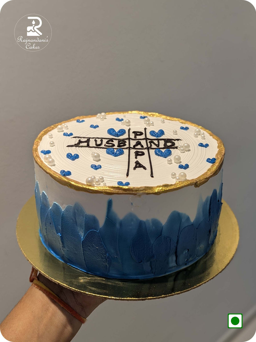 Rajnandani Cakes eggless white and blue whipping cream cake with a gold rim, pearls, and Husband-Papa crossword text.