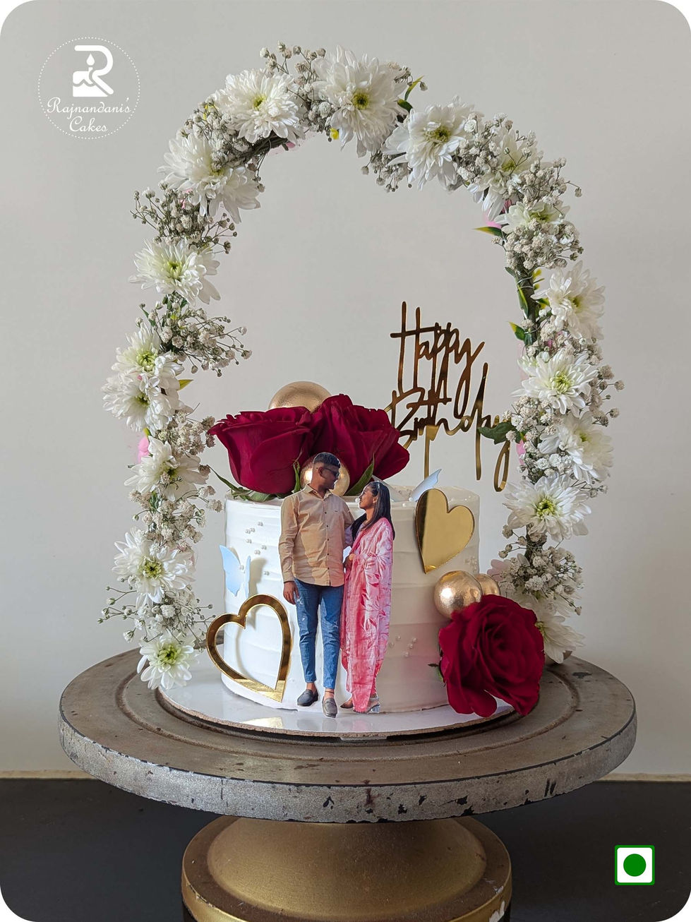 White birthday cake featuring a fresh white flower arch, red roses, golden hearts, and a personalized couple cutout topper.