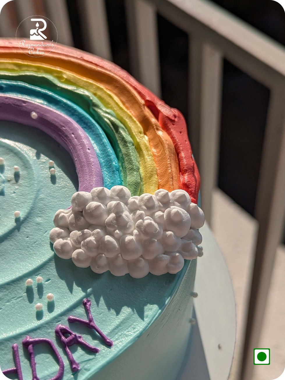 Rainbow first birthday cake featuring piped clouds, sugar pearls, and Happy Birthday lettering on sky blue frosting.