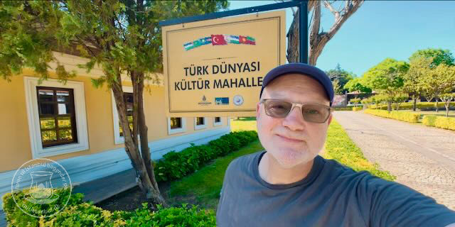 Mand tager selfie foran skiltet TÜRK DÜNYASI KÜLTÜR MAHALLE ved en gul bygning og en vej i solskin.