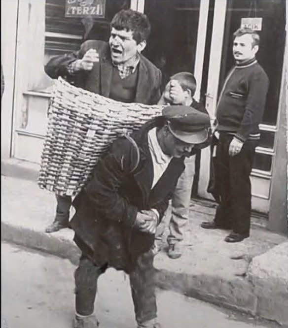 A man shouts while being carried in a large basket on another man's back in a street. A child and a bystander watch. Monochrome scene.
