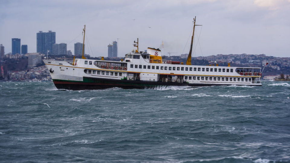 Hvid færge sejler på bølget hav foran byens skyline under grå himmel.