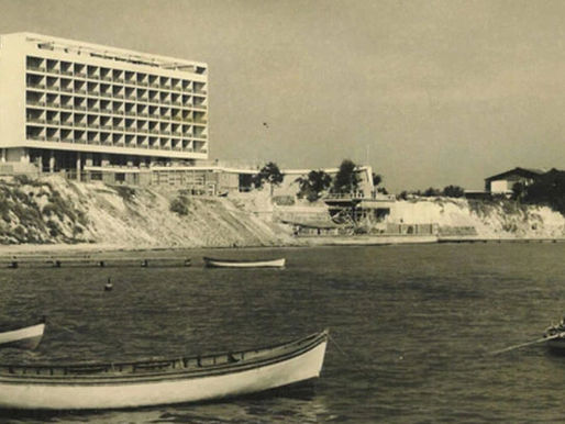 Sort/hvidt foto af hotel ved kysten med både i vandet og en person som ror. Bakken har træer, og der står en fyrtårn. Stemningen er rolig.