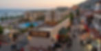 Solnedgang ved Sunset Beach-resort med pool, hav og travl gade med fodgængere, tyrkiske skilte og bakket by.