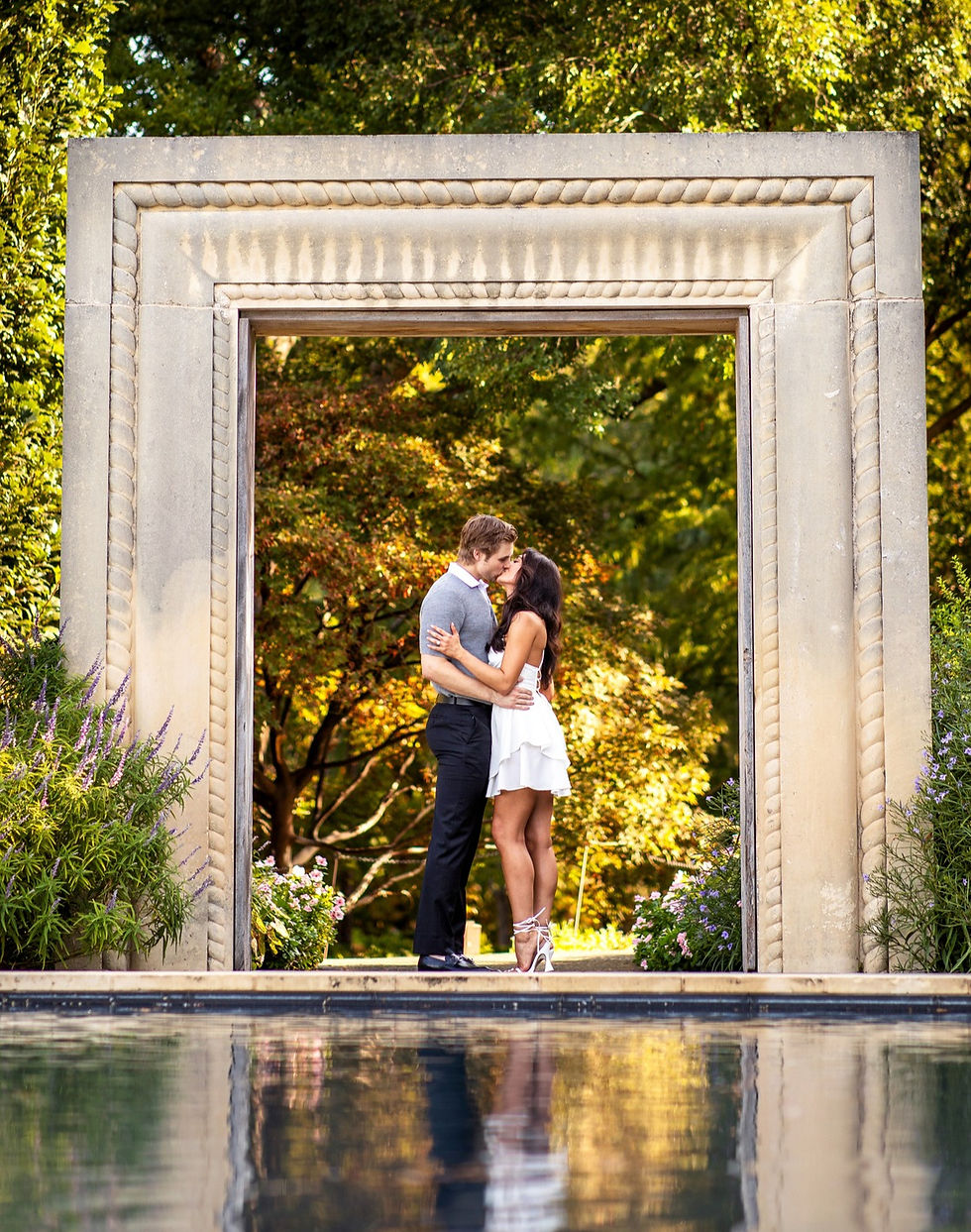 A couple embraces under a stone archway, surrounded by lush greenery. Their reflection appears in a pool, creating a romantic scene.