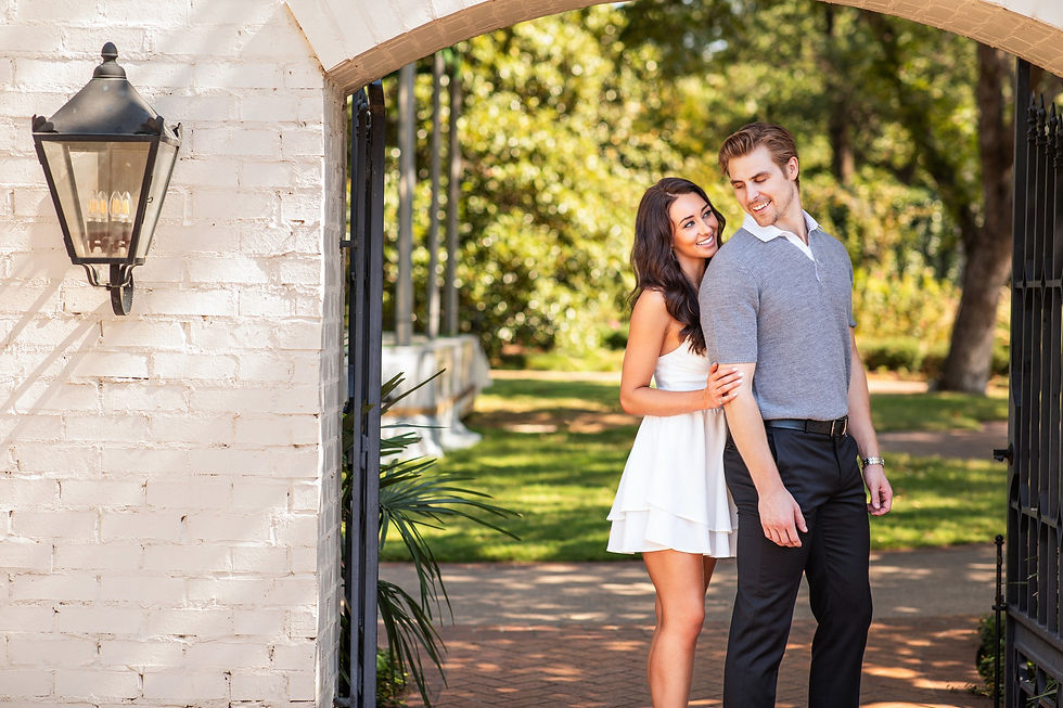 A smiling couple stands in a garden under an archway. She's in a white dress, he's in a gray shirt. A brick wall and lantern are visible.