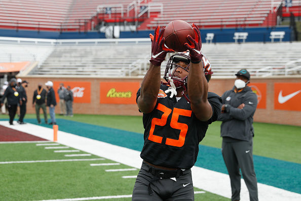 Football player in black jersey, number 25, catching ball on field. Coaches watch nearby. Stadium seats in background, overcast day.