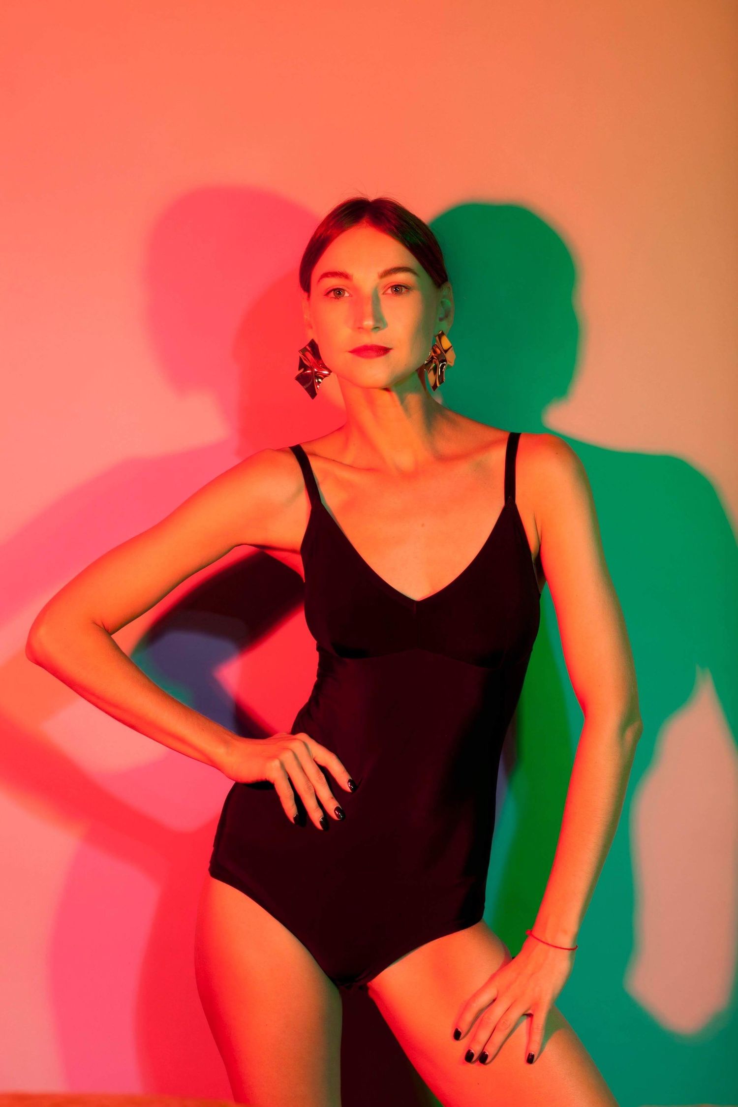 Fashion model in black swimsuit poses against colorful red and green lighting backdrop with dramatic shadows.