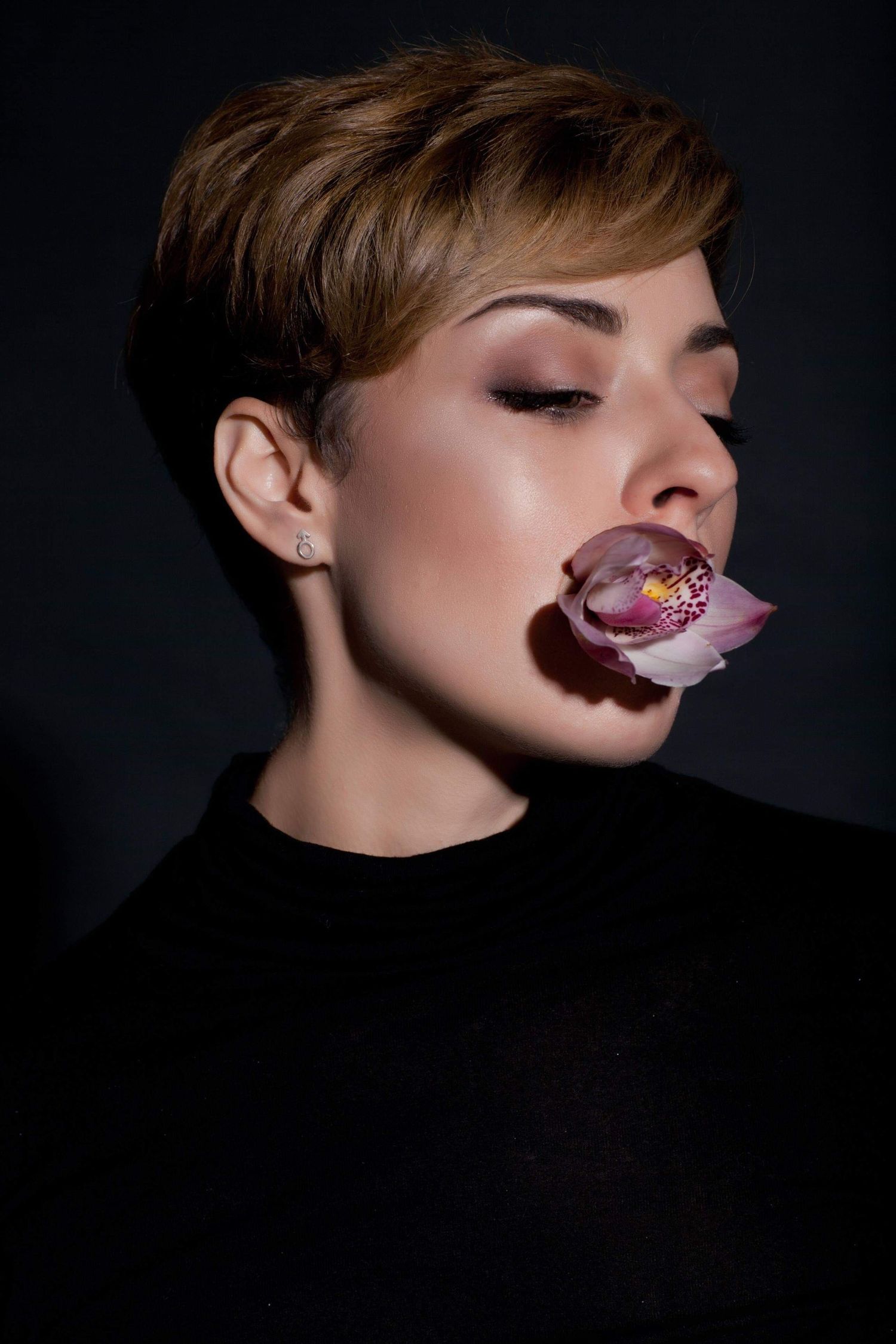 Artistic beauty portrait featuring purple orchid flower placed on dark lips against black background.