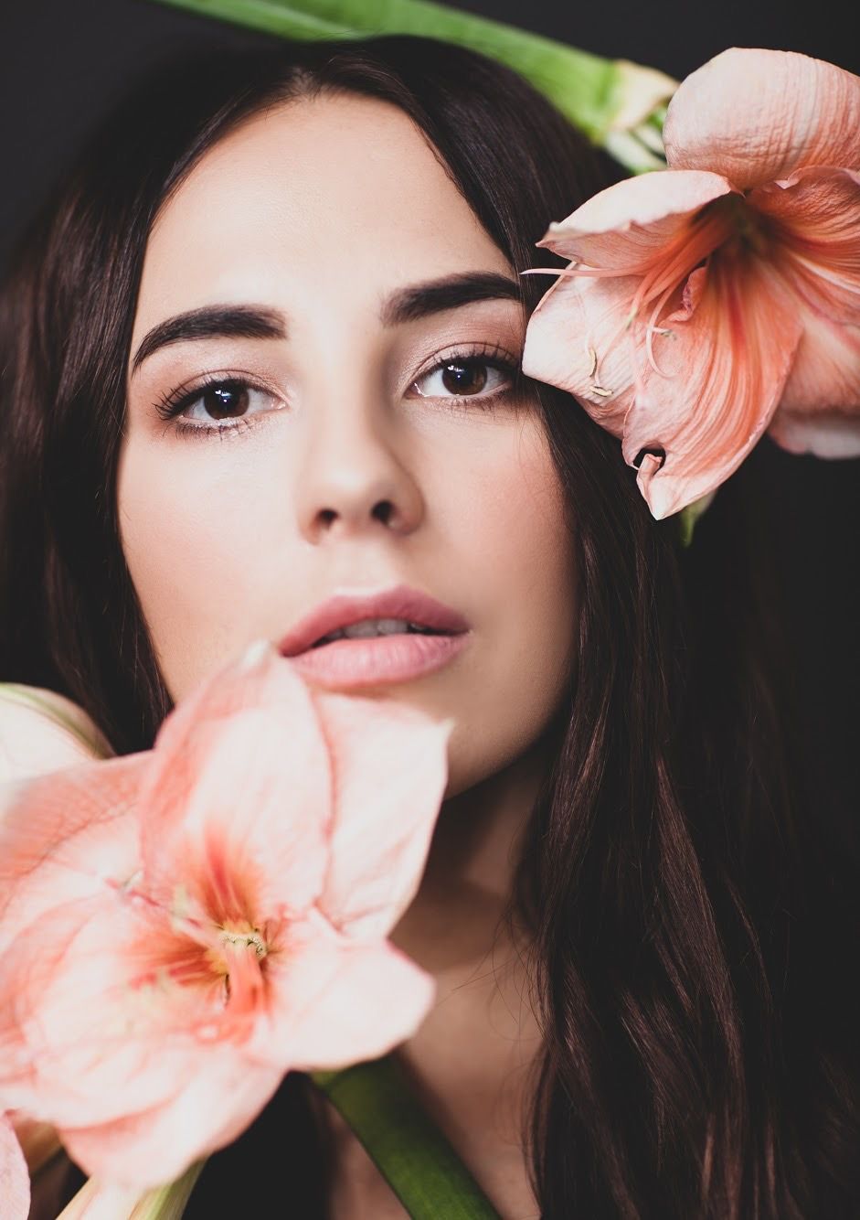 Beauty portrait with soft peach amaryllis flowers framing the face against dark background.