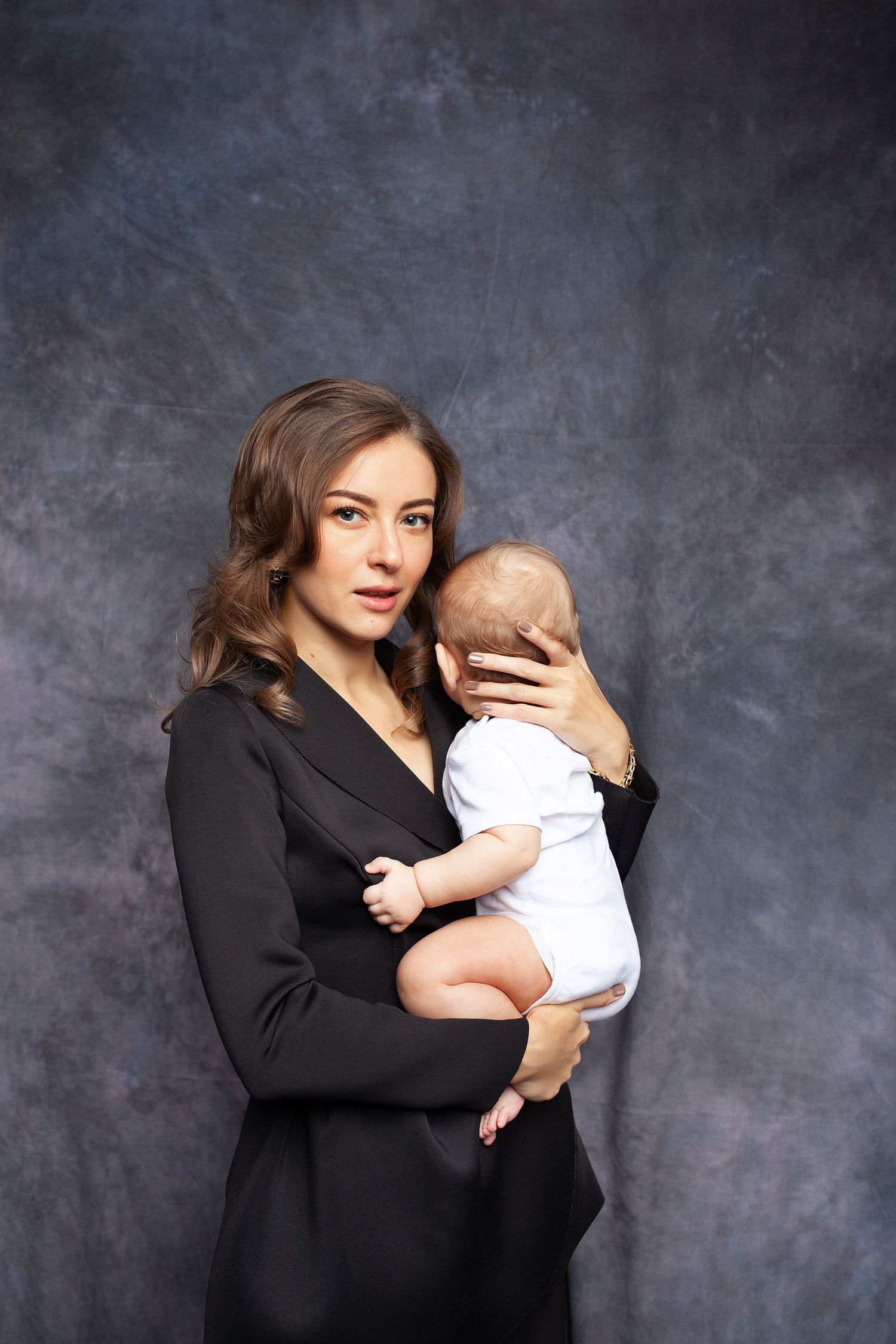 Professional studio portrait of mother in black blazer holding infant in white clothing.