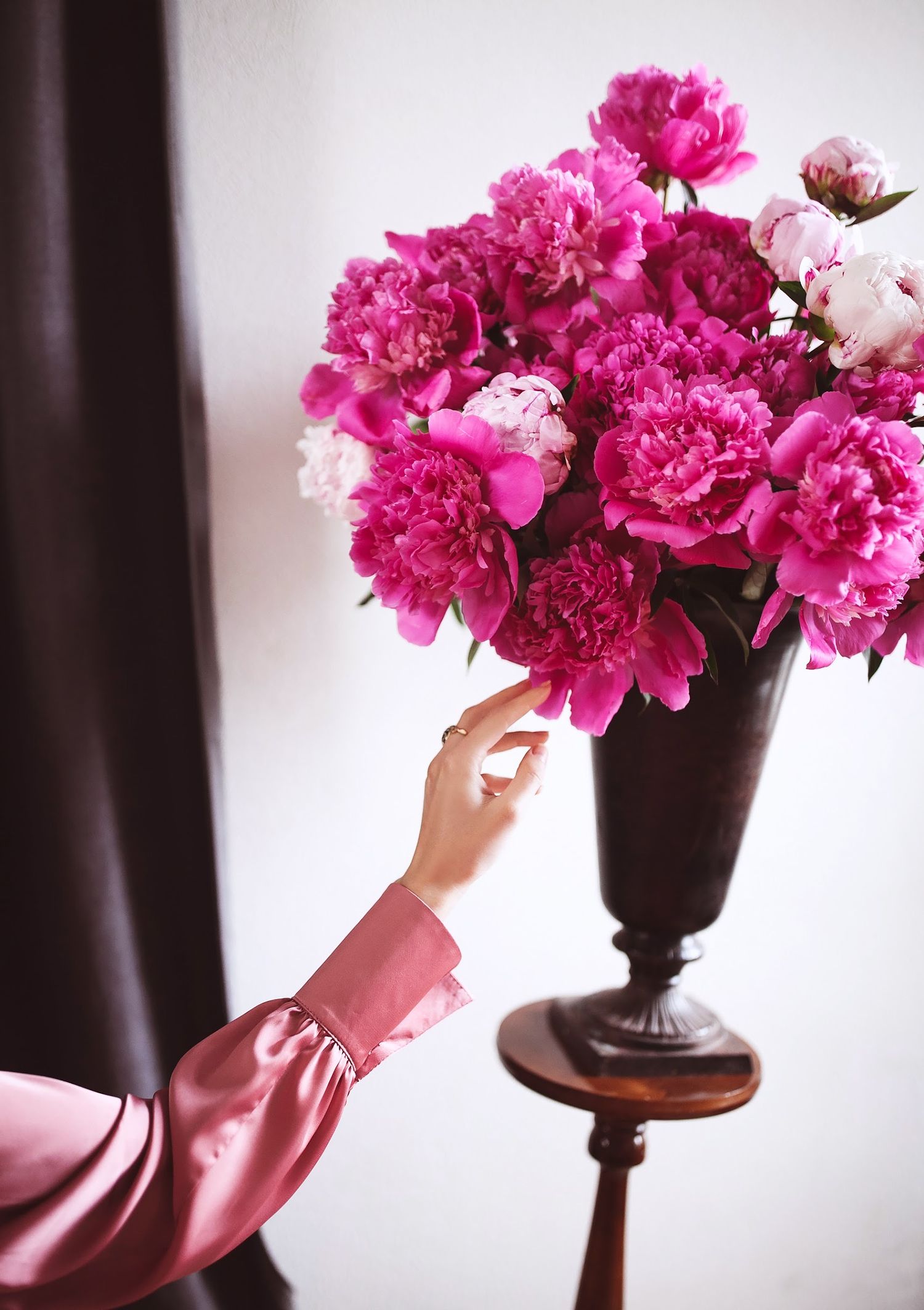 Hand reaching to arrange bright pink peonies in dark vase on wooden stand.