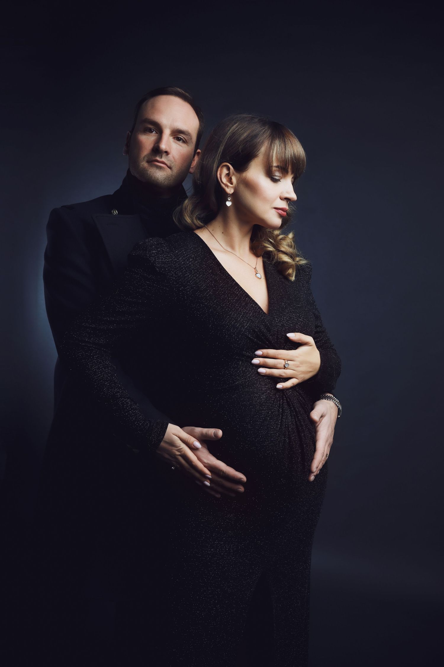 Dramatic couple portrait in black formal wear against dark background.
