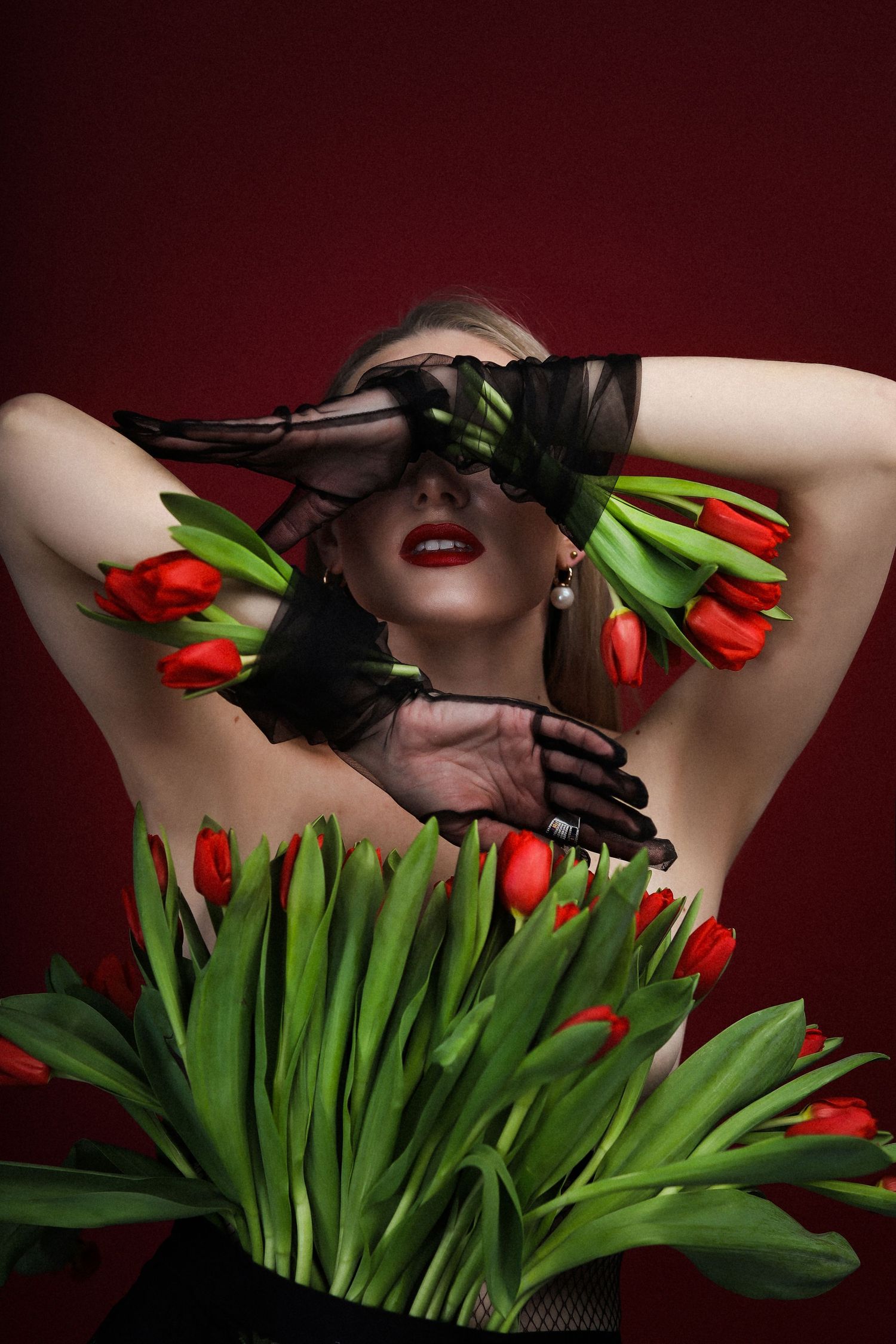 Artistic portrait with red tulips and black lace gloves against a burgundy backdrop.