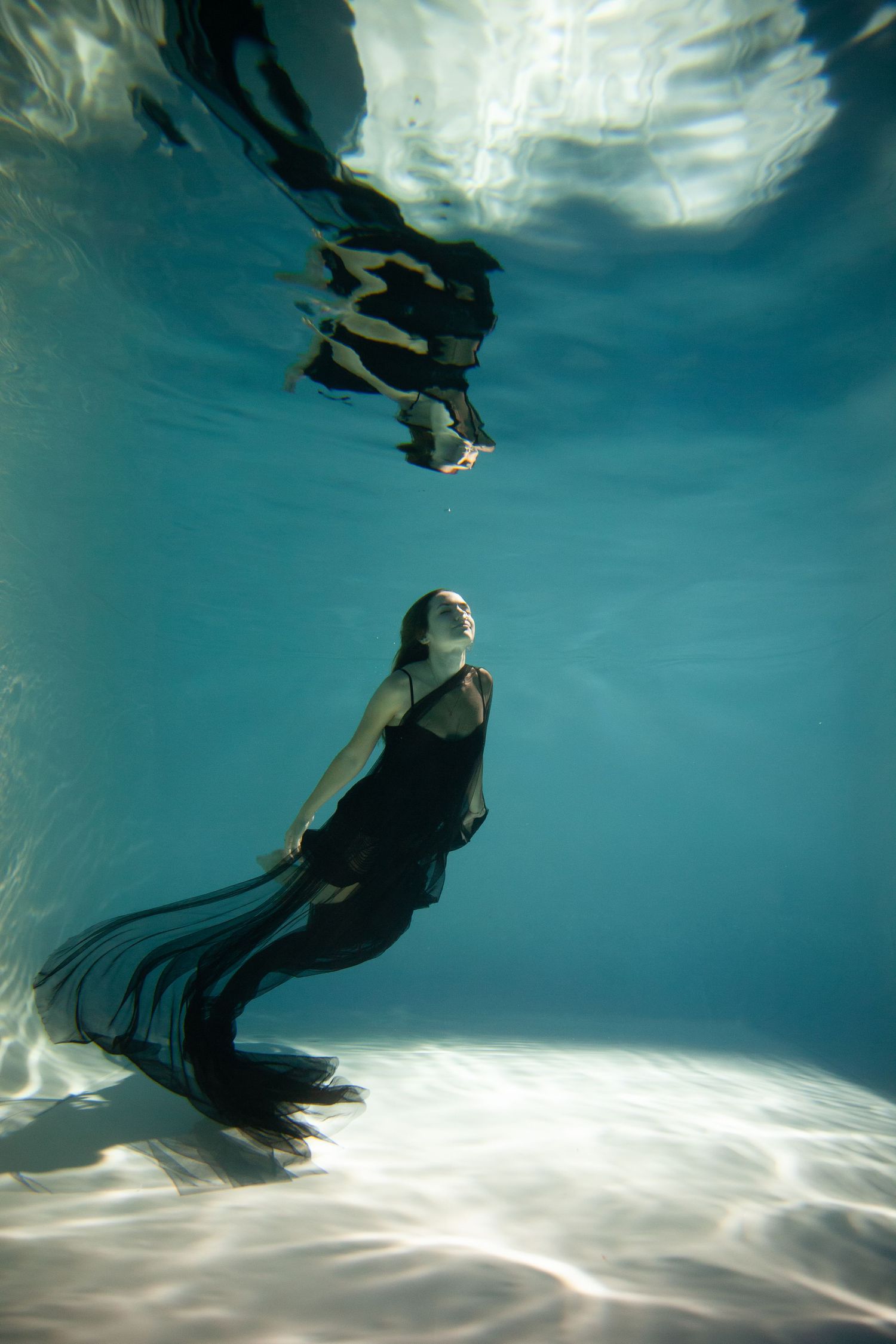 Ethereal underwater portrait of figure in black dress with reflection on surface.