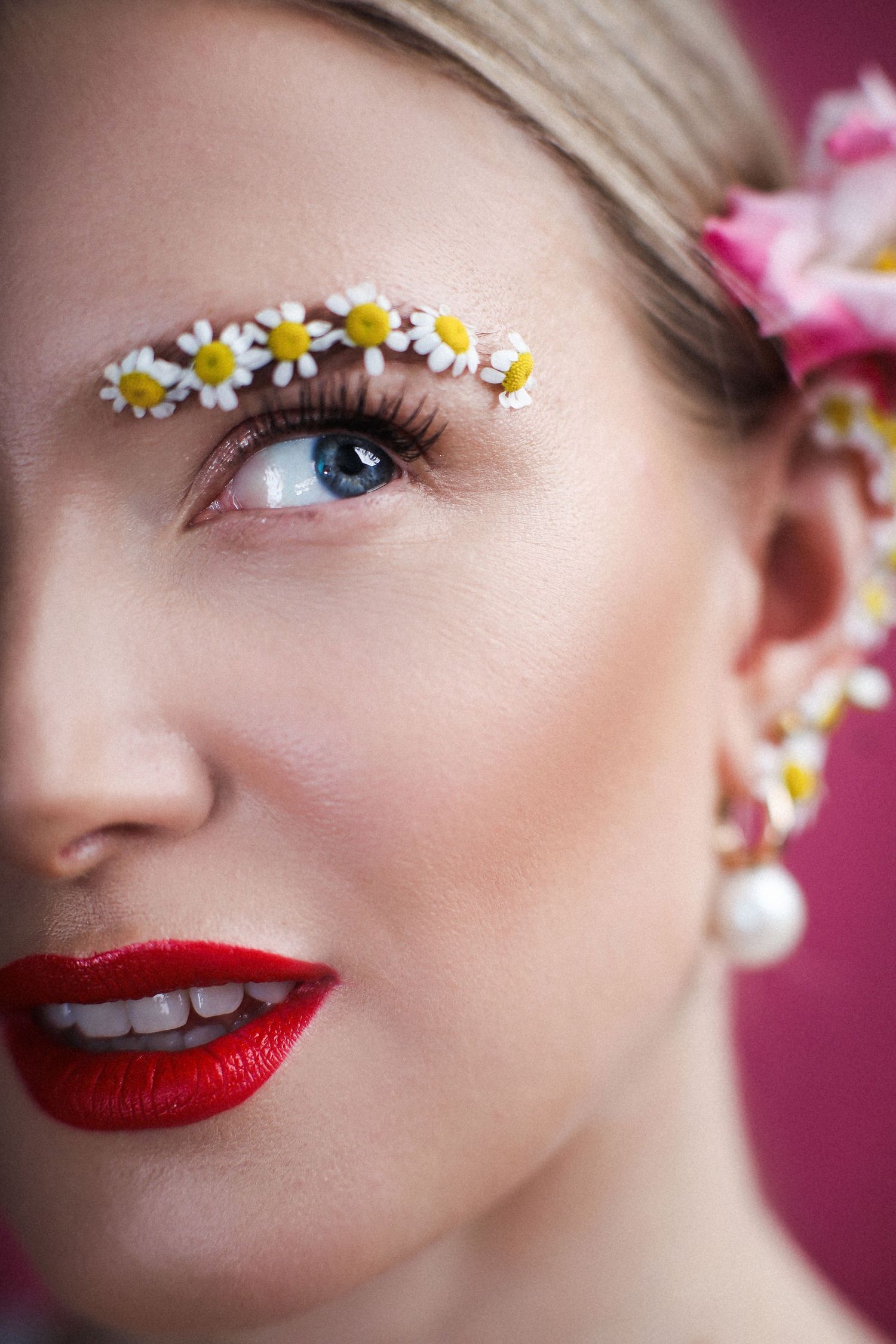 Creative beauty makeup featuring daisy flowers arranged above the eye with bright red lipstick against pink background.
