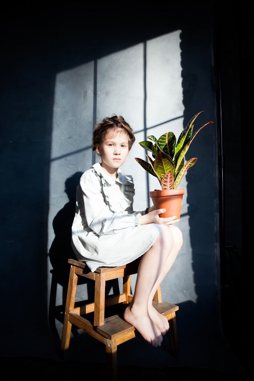 Dramatic portrait of child sitting on wooden stool holding potted plant in sunlit room.