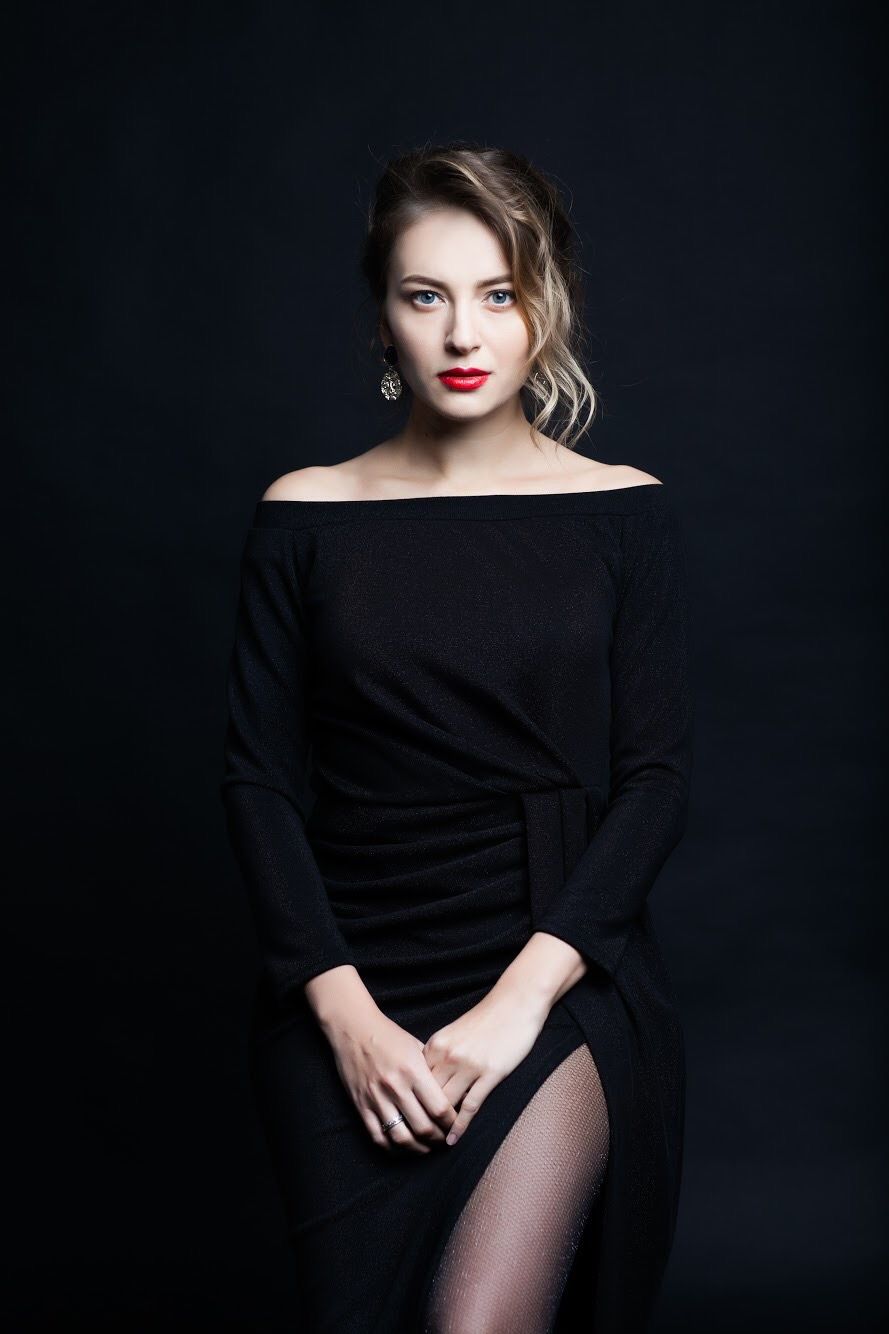 Elegant fashion portrait in black off-shoulder dress against dark backdrop with dramatic lighting.