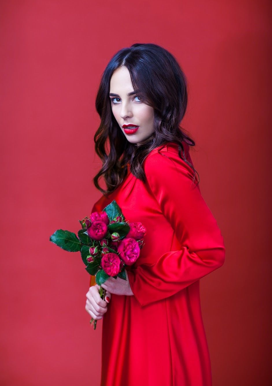Dramatic fashion portrait with red dress and roses against vibrant red background.