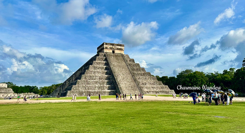 People gather near Chichen Itza pyramid under a blue sky. Lush green grass and trees surround. Text reads "Charmaine Wanders."