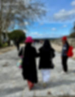 Deux personnes entourées sous un ciel nuageux, une dans un chapeau rose et un sac sculpté. Une troisième personne avec un sac à dos rouge regardant. Les arbres sont bordés sur le chemin.