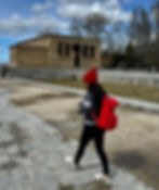 Une personne dans un chapeau rouge et marche sur le dos près d'un vieux bâtiment en pierre. Nuages ciel, arbres nus et chemin pavé.
