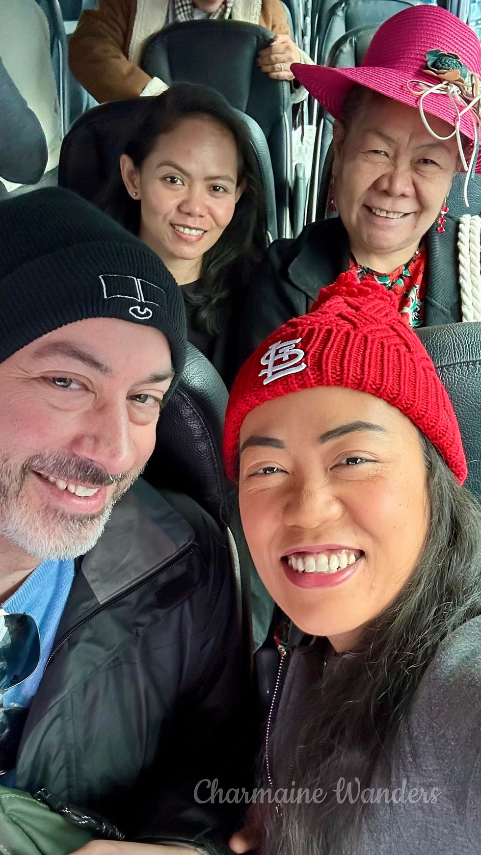 Four people smiling in a bus, wearing colorful hats and beanies. Background shows bus seats. Text: Charmaine Wanders. Mood is joyful.