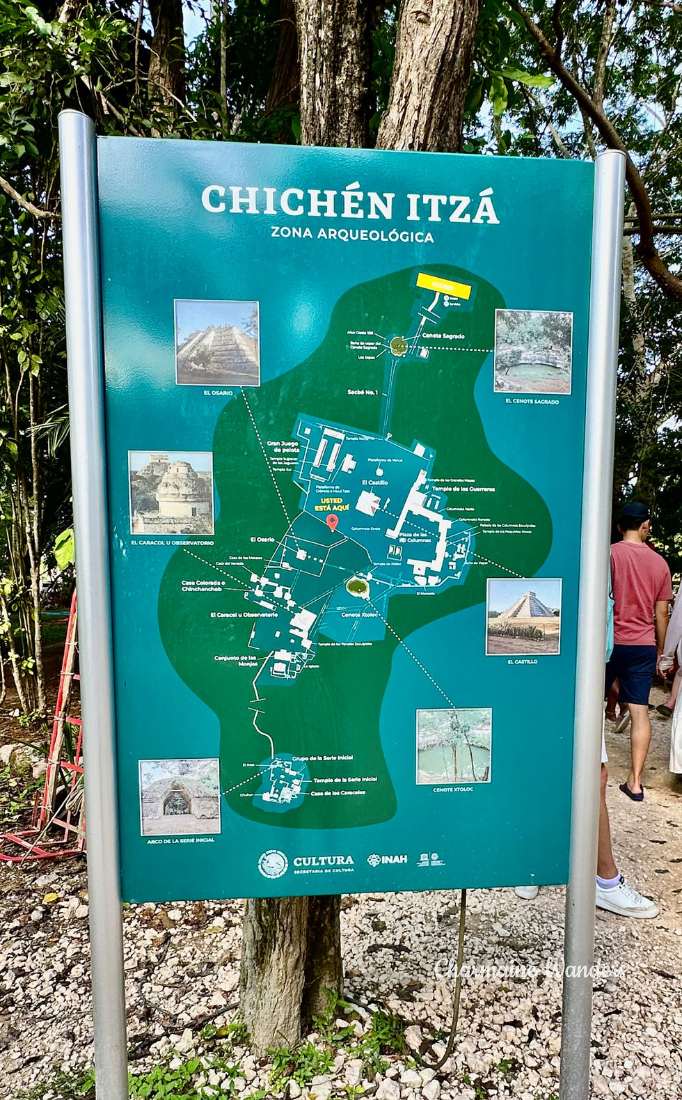 Chichén Itzá map sign with images and labels in a wooded area. Two people partially visible on the right, standing nearby.