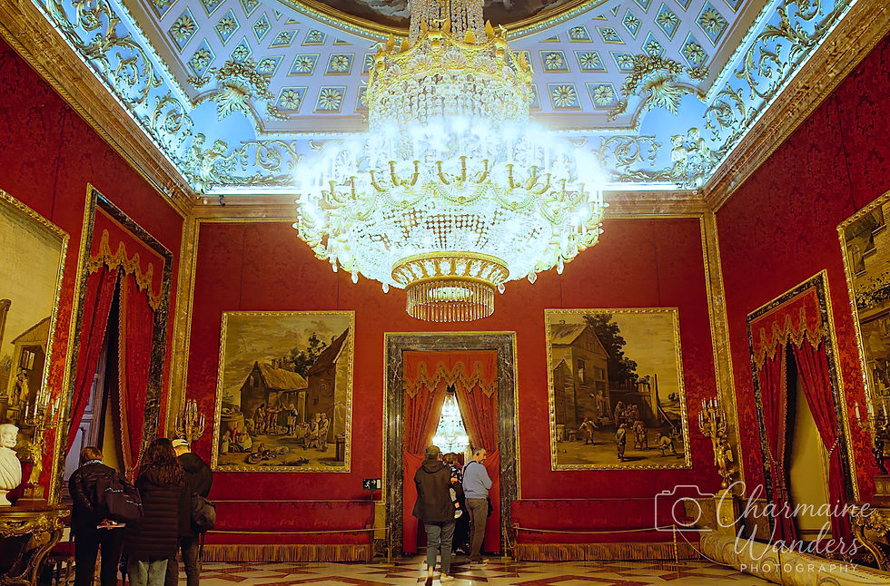 Luxurious room with a grand chandelier, red walls, ornate decor, and large paintings. People observe the artwork and decorations.