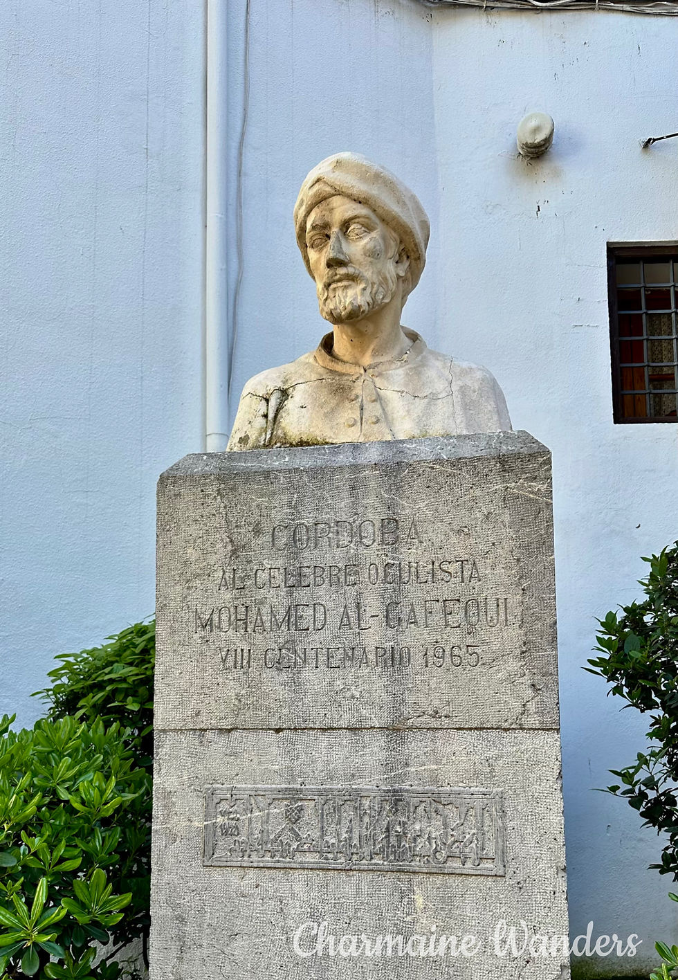 Bust of Mohamed Al-Gafequi on a stone pedestal with inscriptions, set against a pale wall. Green shrubbery accents the sides. Cordoba, Spain. Text: Charmaine Wanders
