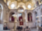 Hall décoratif avec toit arqué, grandes fenêtres et sculptures en marbre. Les gens marchent et admirent l'art. Les rideaux rouges et le logo royal sont visibles.