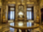 Ornate room with detailed wallpaper, chandeliers, large mirrors, and a reflective table. Elegant chairs and intricate floor patterns. Luxurious ambiance.