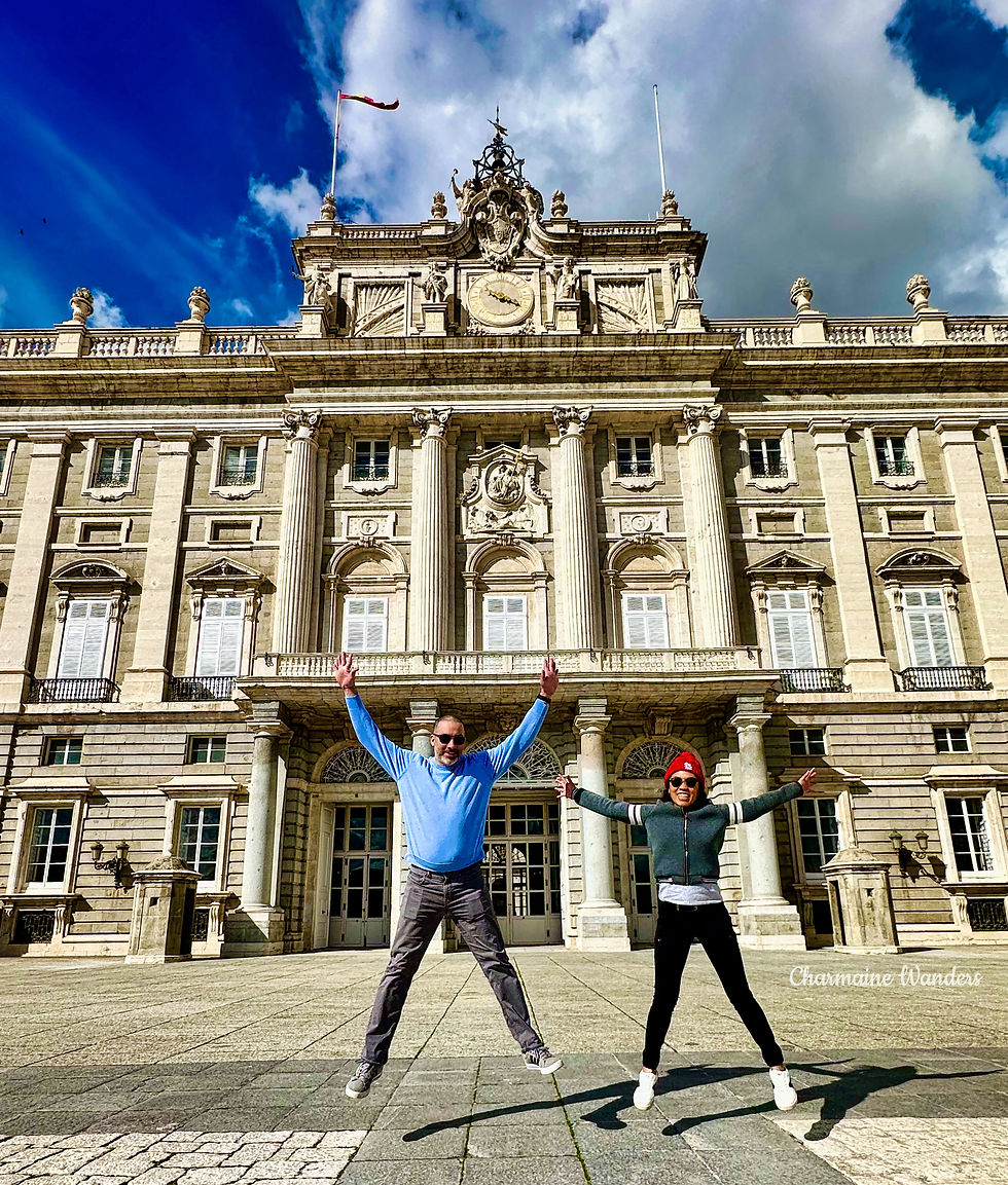 Kevin and Charmaine jump joyfully in front of a grand historic building with ornate facade and columns. Blue sky and clouds above; one wears a red hat.