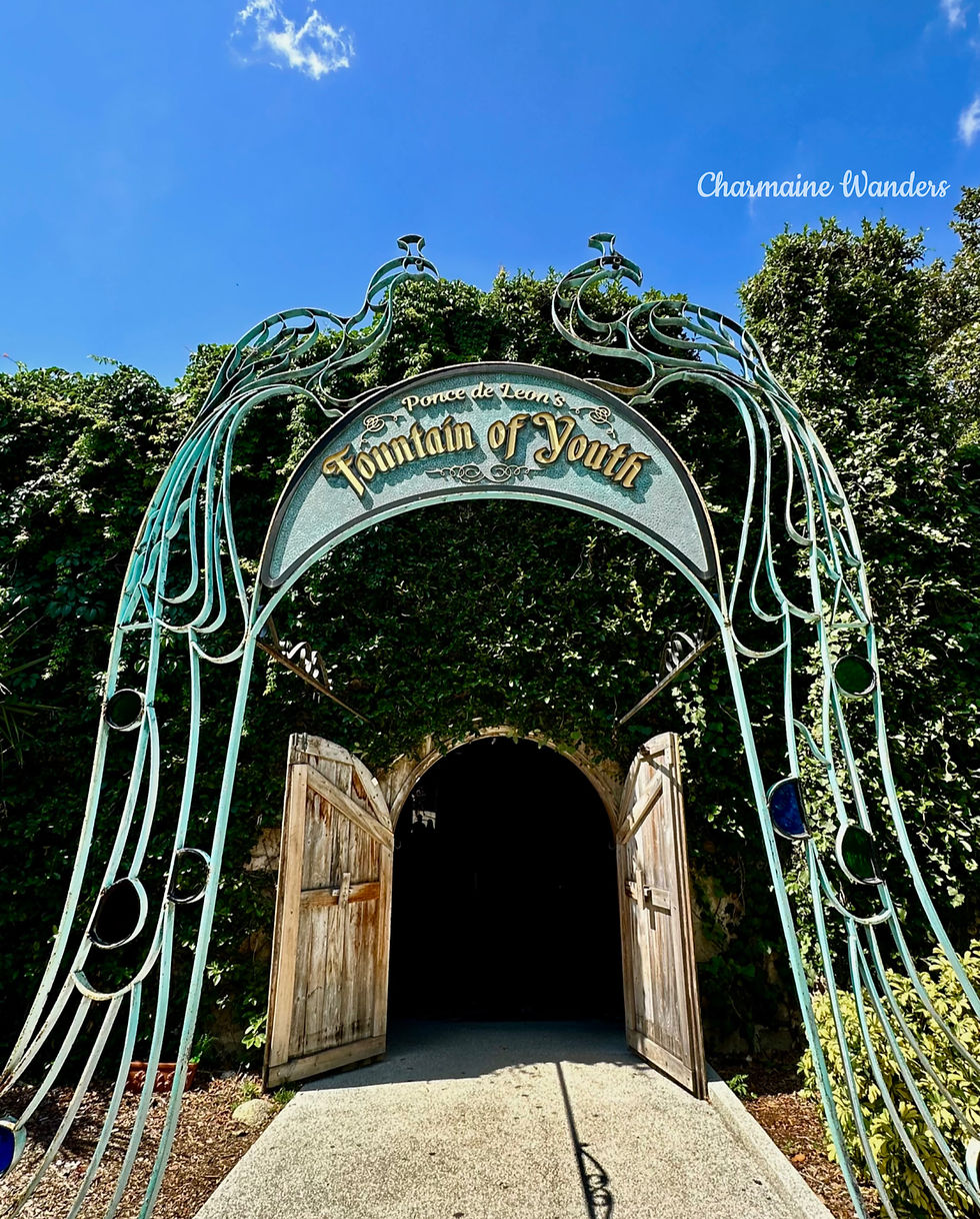 Ornate iron arch reading "Ponce de Leon’s Fountain of Youth" over wooden doors. Lush greenery surrounds, with a bright blue sky above.