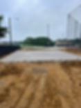 Newly poured concrete slab foundation with dirt foreground, field and building background