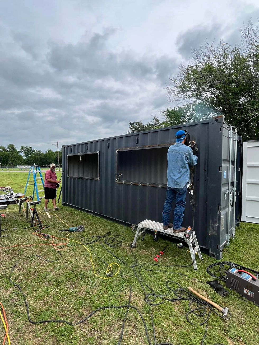 Two workers welding and modifying a dark shipping container outdoors.