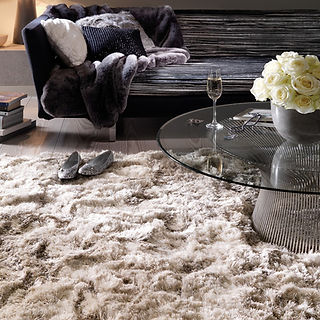 Our very deep shaggy Plush rug in Sand colour, on the floor of a lounge with a sofa and coffee table.