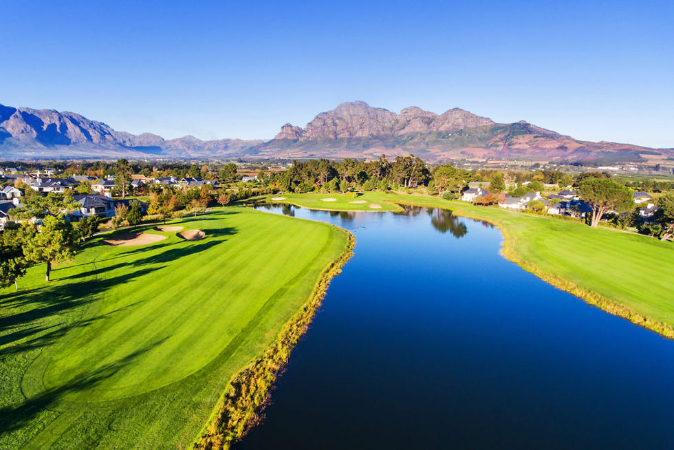 Lush green golf course with a reflective waterway, surrounded by trees and houses, set against a clear blue sky and distant mountains.