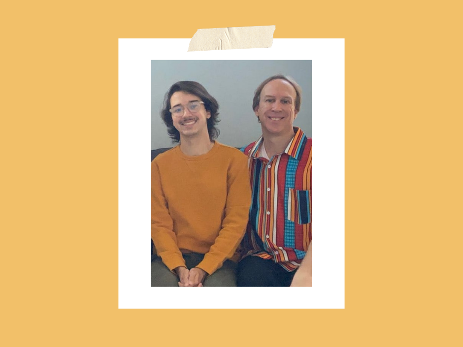 Two smiling people sit indoors; one wears an orange sweater, the other a colorful striped shirt. The background is plain gray.