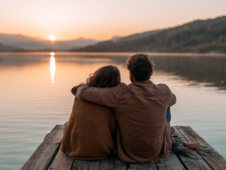 Husband and wife sitting together on a lakeside dock at sunrise, reflecting quietly while overlooking a calm mountain lake and surrounding forest.