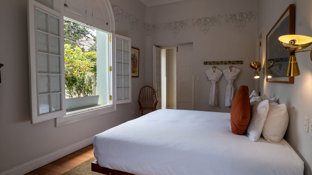 Vue intérieure de la suite de la Villa Barranco avec grand lit, linge blanc, coussins terracotta, appliques dorées et fenêtre ouverte sur jardin.