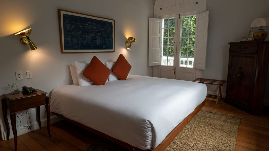 Autre perspective de la suite de la Villa Barranco montrant le lit king size, les tables de nuit en bois, les lampes murales dorées et la fenêtre avec volets.