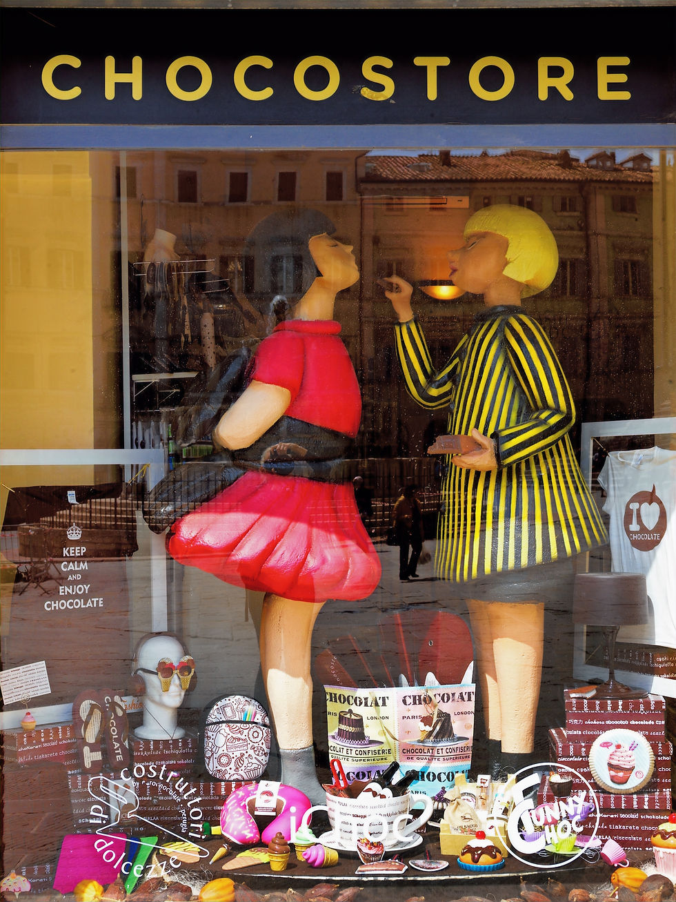 Schaufenster mit zwei bunten Figuren, eine in Rot, die andere in Gelb-Schwarz gestreift. Schokolade und T-Shirts mit Texten im Hintergrund.