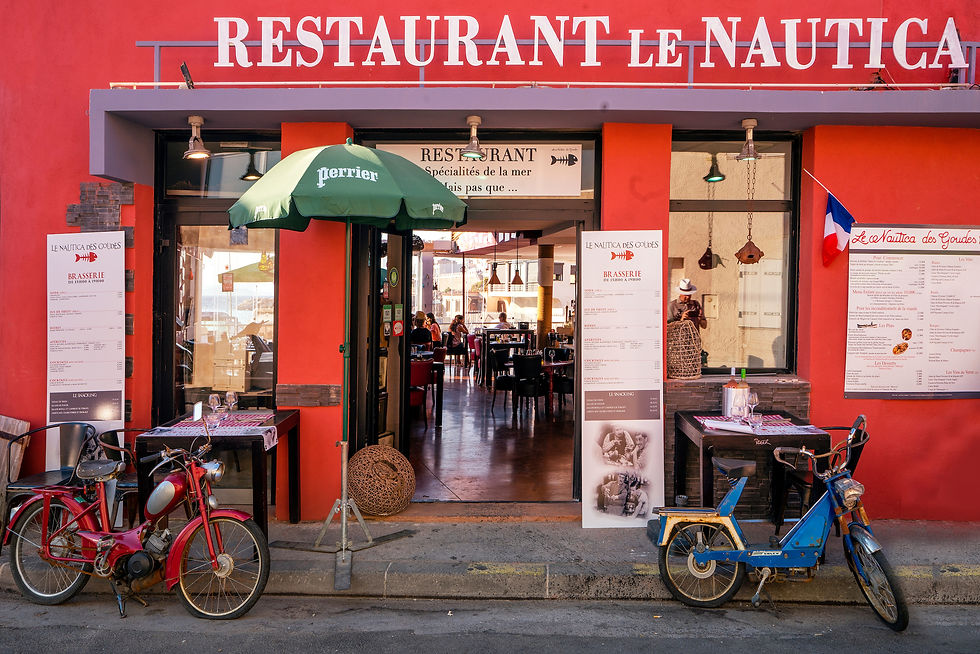 Les Goudes marseille, Rotes Restaurant mit Inschrift "RESTAURANT LE NAUTICA", zwei Mopeds vorne. Grüner Sonnenschirm, Menütafeln, Menschen im Inneren. Französische Flagge.