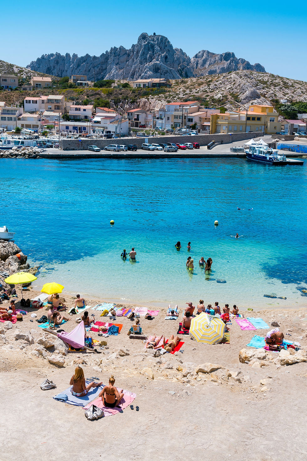 Menschen genießen einen sonnigen Strand; einige schwimmen im klaren blauen Wasser. Im Hintergrund sind Häuser und felsige Hügel zu sehen.