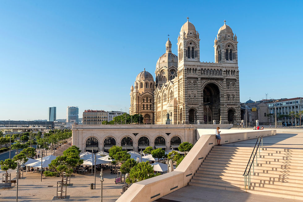 Kathedrale in Marseille bei klarem Himmel. Menschen spazieren auf breiter Treppe. Moderne und historische Gebäude im Hintergrund.