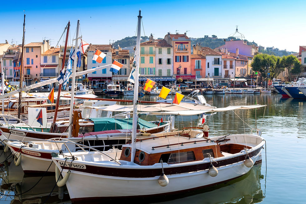 Boote im malerischen Hafen von Cassis, vor bunten Häusern. Bunte Flaggen schmücken die Boote, Sonnenschein reflektiert im Wasser. Entspannte Stimmung.