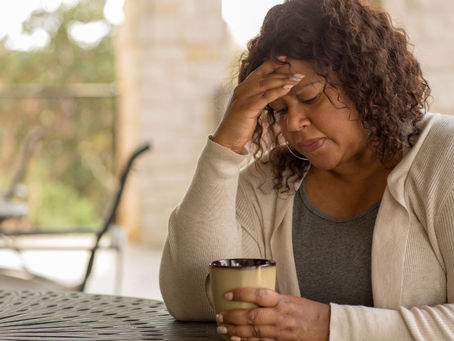 A woman with curly hair holds a mug, resting her head in her hand, looking pensive. She's seated at a table outdoors, with blurred greenery.