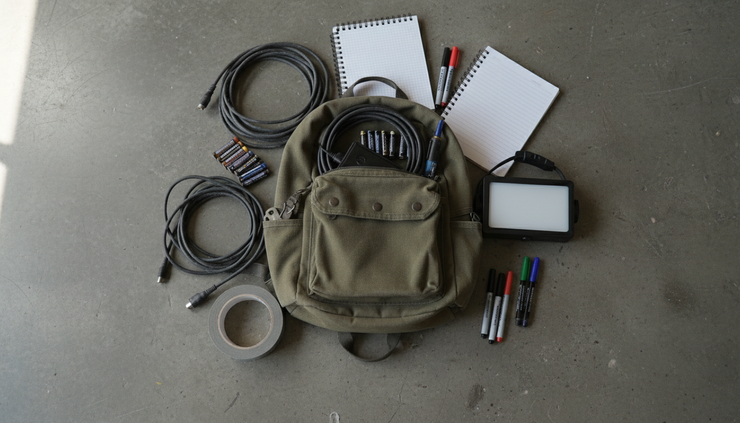Overhead view of a small backpack filled with batteries, tape, notebooks, and cables